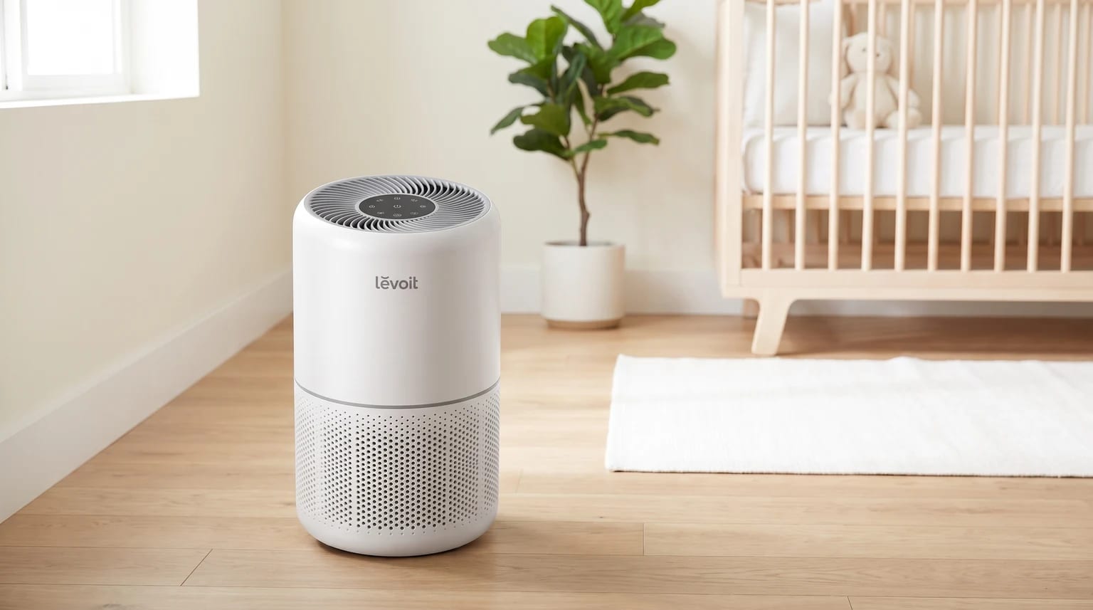 Un purificador de aire Levoit blanco en el suelo de madera de la habitación de un bebé, con una cuna y una planta al fondo.