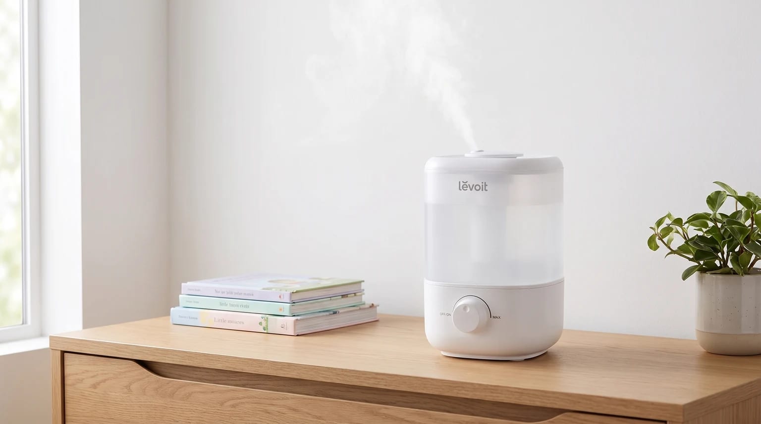 Un humidificador blanco Levoit expulsa vapor frío sobre una cómoda de madera, junto a una pila de libros infantiles y una planta.
