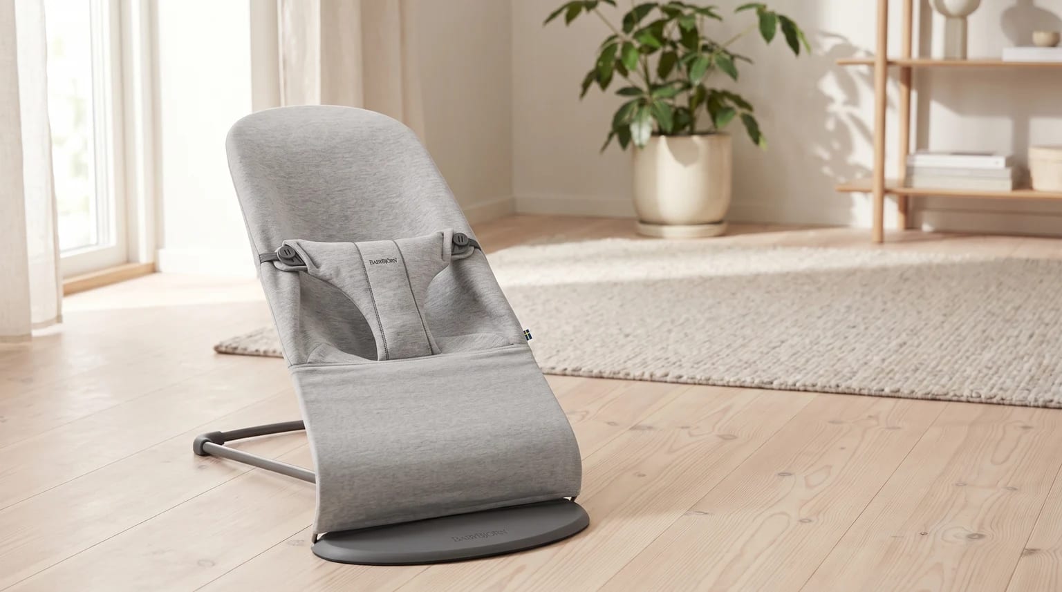 Hamaca para bebé de BabyBjörn en tela gris claro, sobre un suelo de madera en una habitación luminosa.