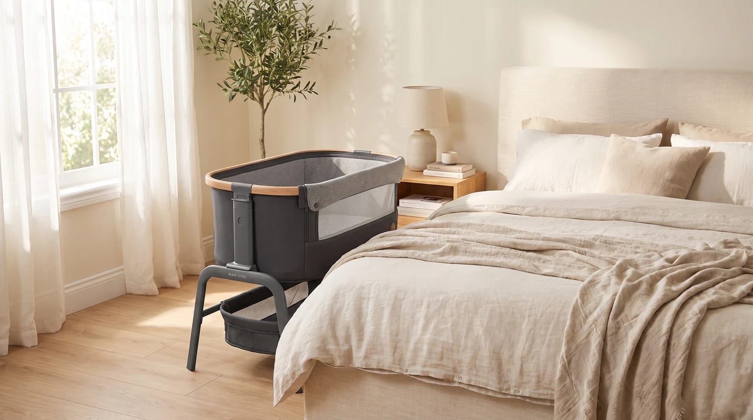 Una minicuna de colecho gris con borde de madera, colocada junto a una cama con ropa de lino en un dormitorio luminoso y tranquilo.