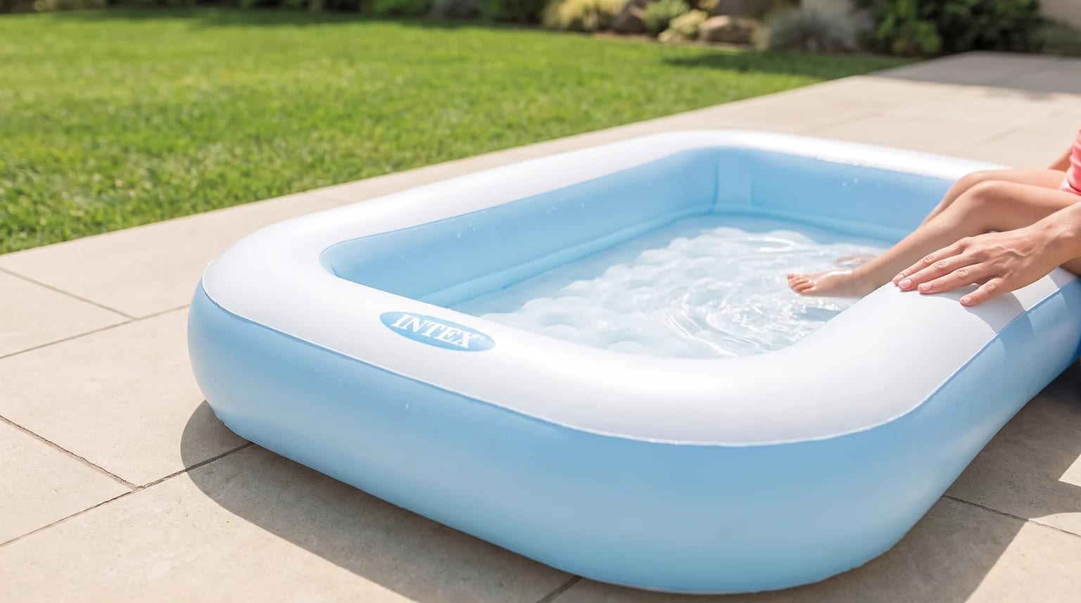 Piscina infantil hinchable azul y blanca en un patio, con una persona adulta sentada al borde y los pies en el agua.