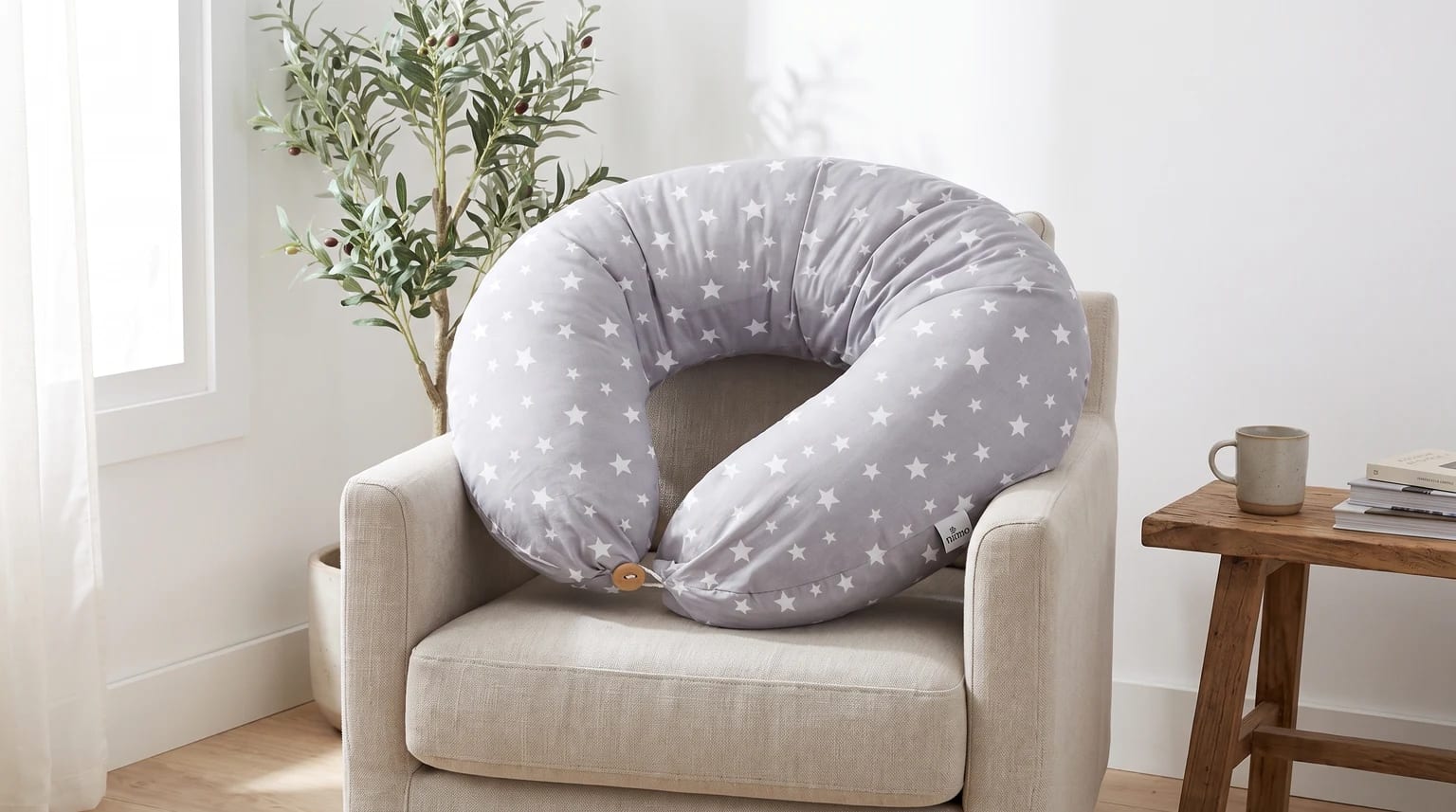 Un cojín de lactancia gris con estampado de estrellas blancas descansa sobre un sillón de tela en una habitación luminosa.