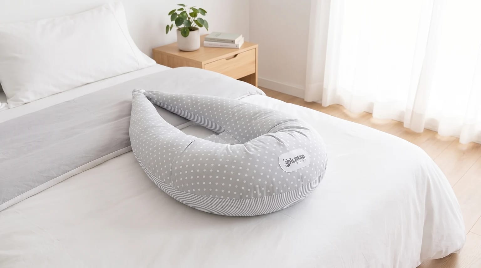 Almohada de embarazo semicircular gris con estampado de lunares y rayas, colocada sobre una cama con sábanas blancas en un dormitorio luminoso.