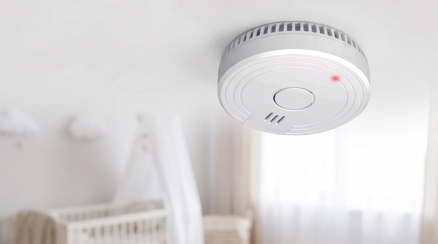 Un detector de humo blanco instalado en el techo de una habitación de bebé, con una cuna desenfocada al fondo.