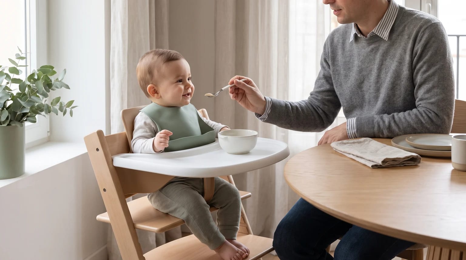 Un padre sentado a una mesa de madera le da de comer con una cuchara a un bebé sonriente que está en una trona.