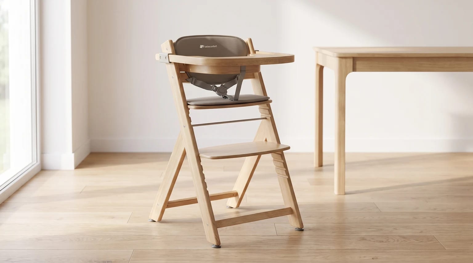 Una trona evolutiva de madera clara con cojines color topo y bandeja, colocada en un comedor luminoso con suelo de parqué.