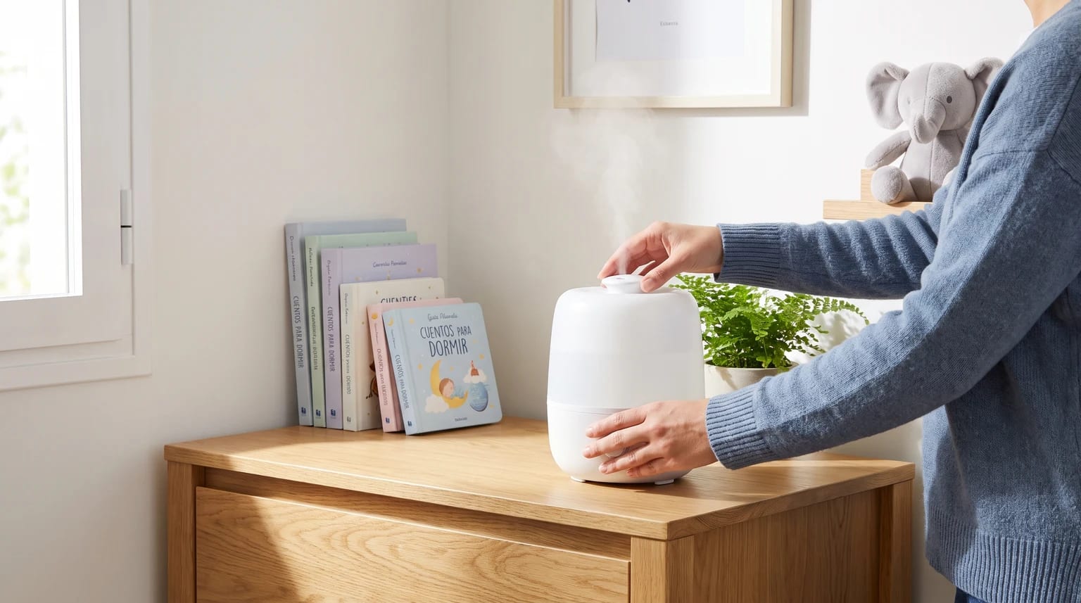 Una persona con un jersey azul ajusta un humidificador blanco sobre una cómoda de madera, junto a unos libros infantiles.
